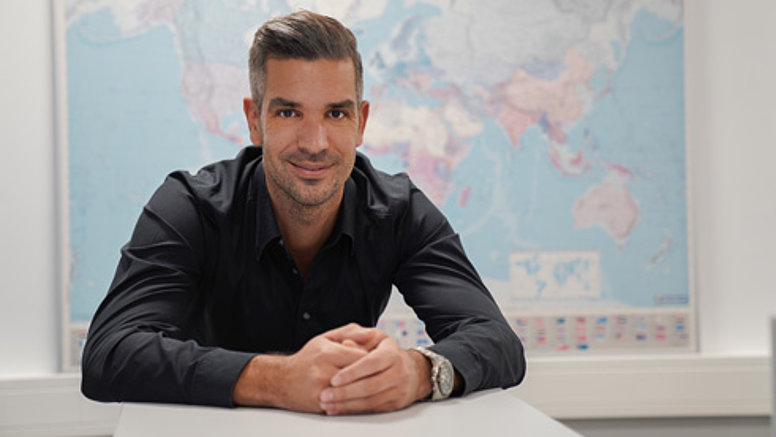 We are international - Our values We are international - employee sits at desk and smiles, in the background is a world map.